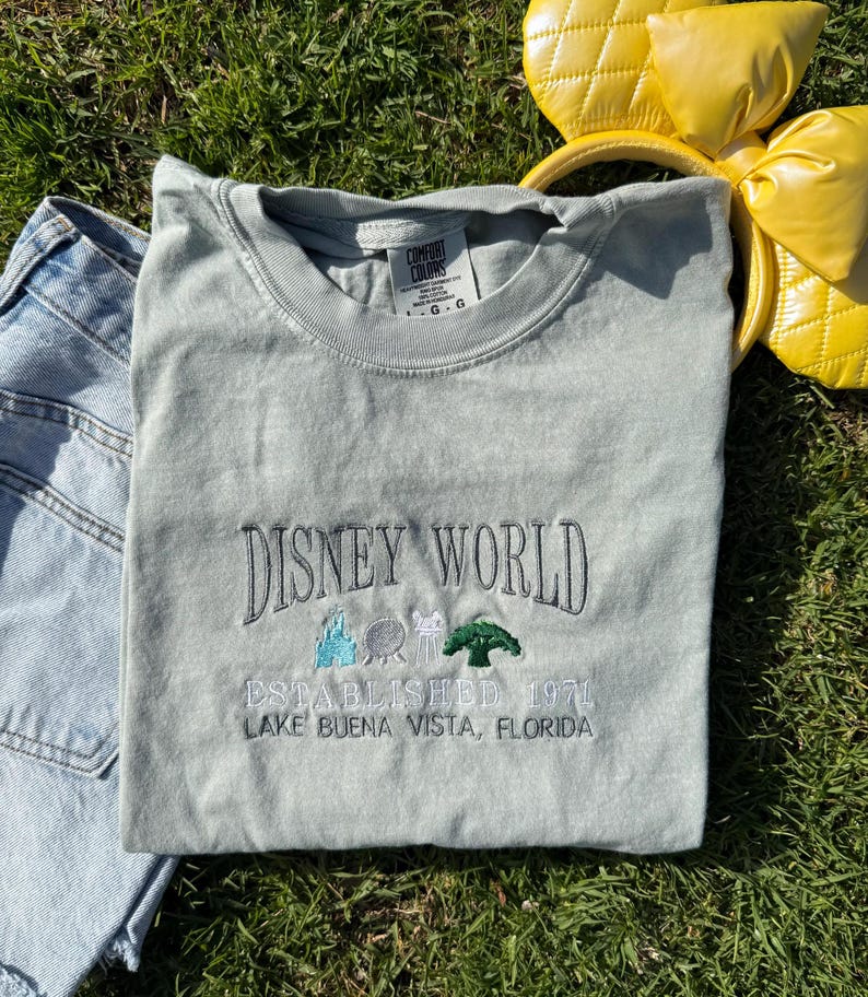 Puede incluir: Una camiseta gris claro con el texto "DISNEY WORLD ESTABLISHED 1971 LAKE BUENA VISTA, FLORIDA" bordado en la parte delantera. El texto est&aacute; en negro con un &aacute;rbol verde, un castillo azul, una bola gris y una cabeza de Mickey Mouse blanca bordada encima.