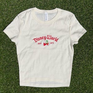 K&ouml;nnte beinhalten: Cremefarbenes Crop-Top mit dem Schriftzug "Disney World" in roter Schrift, zusammen mit "EST. 1971" und einer Kirschgrafik. Das Shirt ist aus weichem Material und hat einen Rundhalsausschnitt.