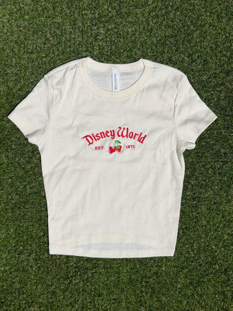 K&ouml;nnte beinhalten: Cremefarbenes Crop-Top mit dem Schriftzug "Disney World" in roter Schrift, zusammen mit "EST. 1971" und einer Kirschgrafik. Das Shirt ist aus weichem Material und hat einen Rundhalsausschnitt.
