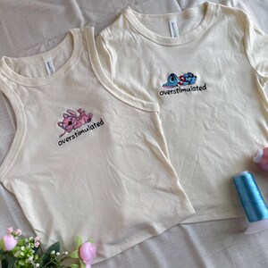 May include: Two cream-colored t-shirts with embroidered cartoon characters and the word "overstimulated." One is a tank top, the other a short-sleeved crew neck. Pink and blue thread spools and faux flowers are in the background.