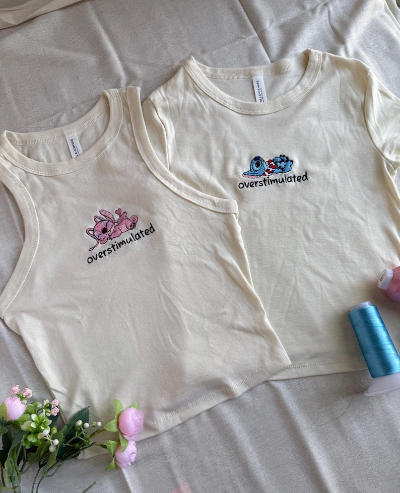 May include: Two cream-colored t-shirts with embroidered cartoon characters and the word "overstimulated." One is a tank top, the other a short-sleeved crew neck. Pink and blue thread spools and faux flowers are in the background.