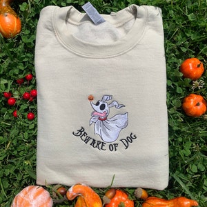 Puede incluir: Una sudadera beige con una imagen bordada de Zero, el perro fantasma de Pesadilla antes de Navidad, sosteniendo una calabaza tallada. El texto "Beware of Dog" est&aacute; bordado debajo de la imagen.