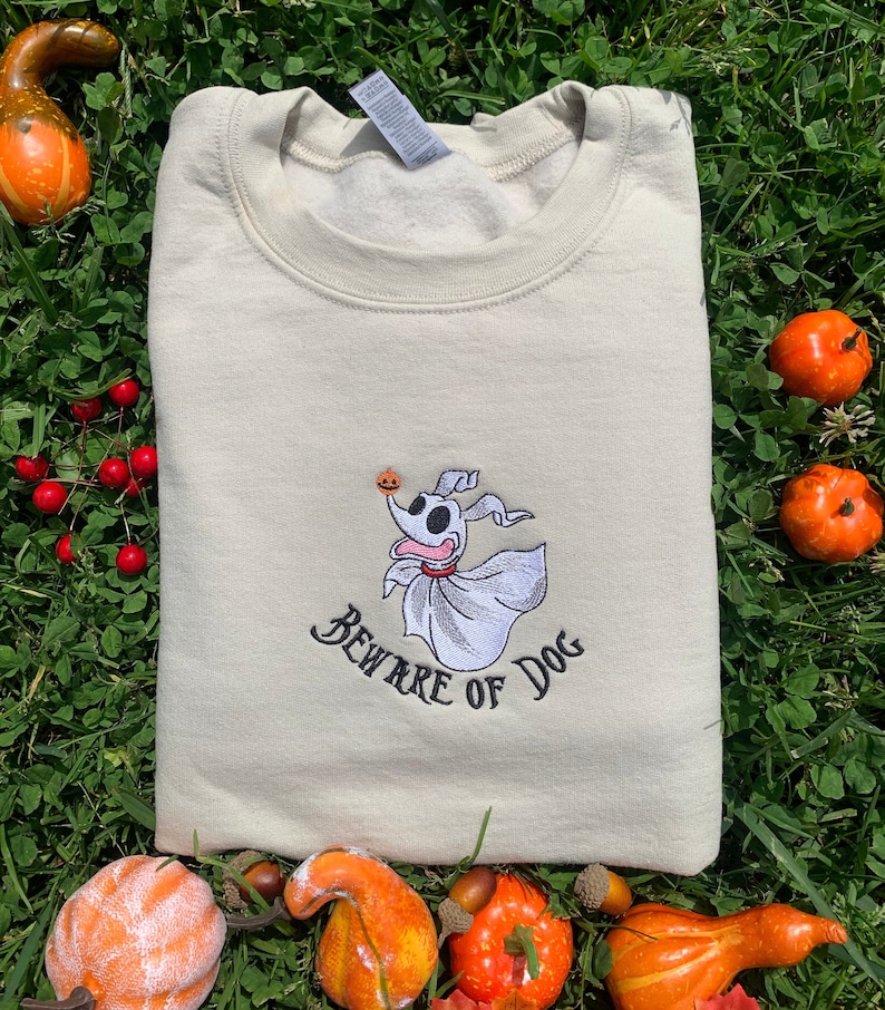 Puede incluir: Una sudadera beige con una imagen bordada de Zero, el perro fantasma de Pesadilla antes de Navidad, sosteniendo una calabaza tallada. El texto "Beware of Dog" est&aacute; bordado debajo de la imagen.