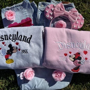 May include: Two sweatshirts, one gray and one pink, embroidered with "Disneyland" and the year "1955", featuring Mickey and Minnie Mouse. A pink headband with a bow and pearl accents, and two pairs of denim jeans are also in the image. Pink roses are scattered around.