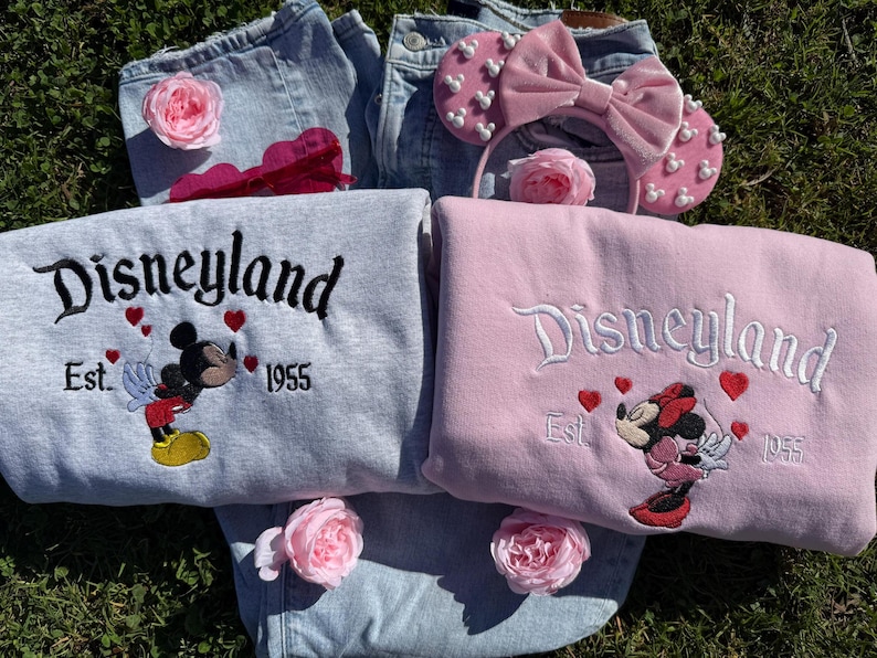 May include: Two sweatshirts, one gray and one pink, embroidered with "Disneyland" and the year "1955", featuring Mickey and Minnie Mouse. A pink headband with a bow and pearl accents, and two pairs of denim jeans are also in the image. Pink roses are scattered around.