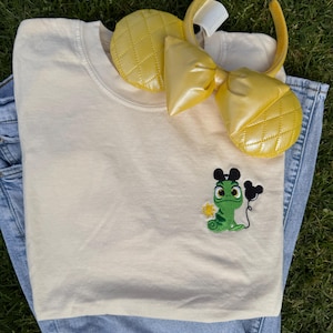 May include: Cream-colored t-shirt with an embroidered green chameleon holding a black balloon. A pair of yellow quilted ears with a matching bow are on top of the shirt. The shirt is on top of a pair of blue jeans.