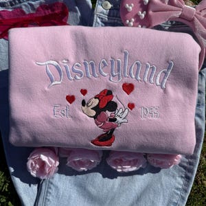 May include: A light pink Disneyland sweatshirt with white embroidered lettering and a Minnie Mouse graphic. The sweatshirt features red heart accents and the text "Est. 1955". The sweatshirt is folded on top of blue denim jeans.