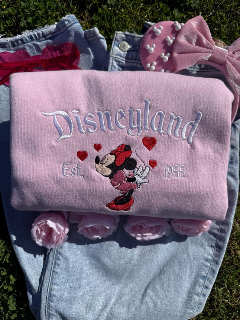 May include: A light pink Disneyland sweatshirt with white embroidered lettering and a Minnie Mouse graphic. The sweatshirt features red heart accents and the text "Est. 1955". The sweatshirt is folded on top of blue denim jeans.