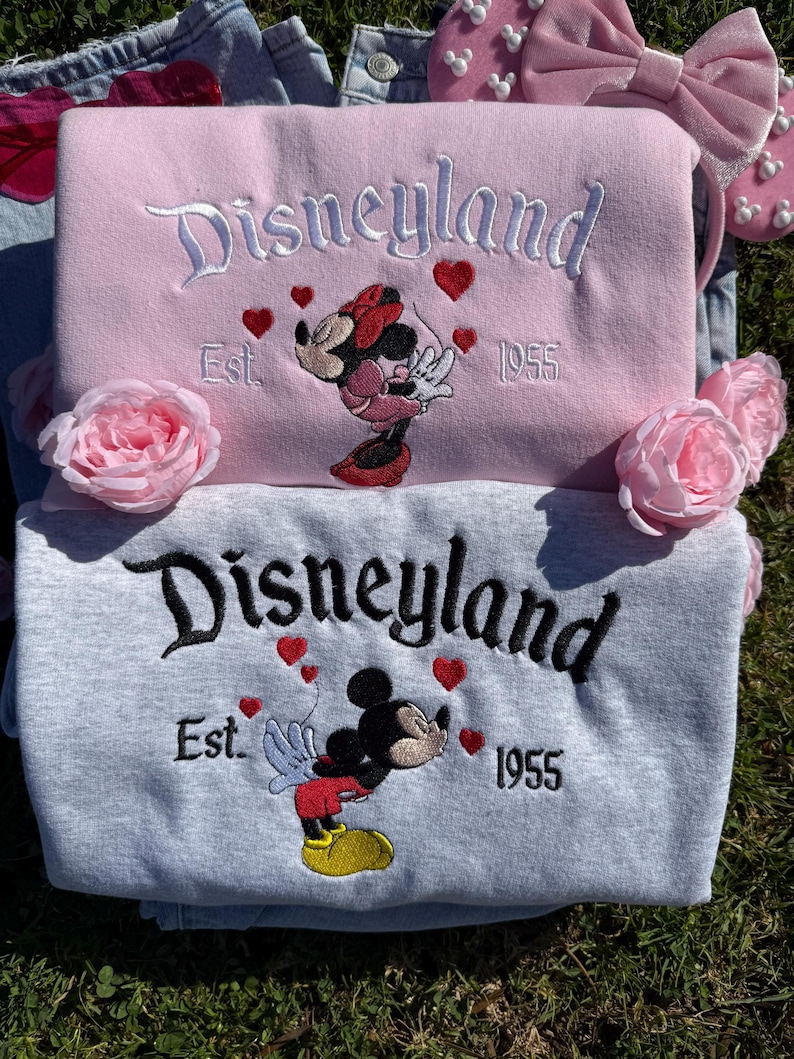 May include: Two sweatshirts, one pink and one gray, embroidered with "Disneyland" in script. The pink sweatshirt features Minnie Mouse, the gray one Mickey Mouse, both with hearts and "Est. 1955." Pink rose accents and a pink bow headband are also visible.
