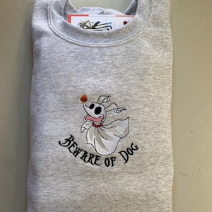 Puede incluir: Una sudadera gris con una imagen bordada de un perro fantasma con el texto "Beware of Dog" debajo. El perro fantasma sostiene una peque&ntilde;a calabaza naranja.