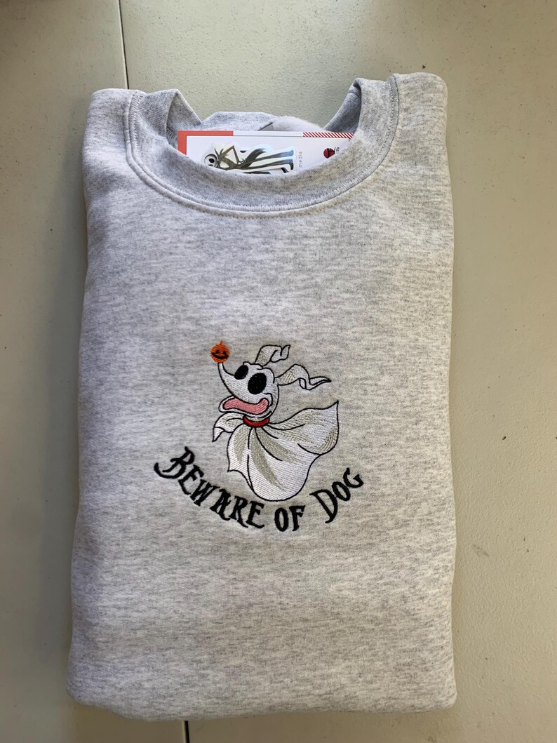 Puede incluir: Una sudadera gris con una imagen bordada de un perro fantasma con el texto "Beware of Dog" debajo. El perro fantasma sostiene una peque&ntilde;a calabaza naranja.