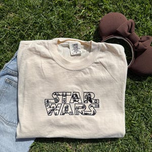 May include: A beige t-shirt with a black "Star Wars" graphic. The graphic is made up of various Star Wars characters and symbols. The t-shirt has a tag that says "Comfort Colors" and "L-G-G".