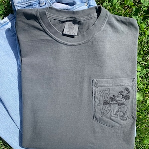 May include: A dark grey t-shirt with a pocket featuring an embroidered image of Mickey Mouse holding a ship's wheel. The t-shirt has a Comfort Colors tag.
