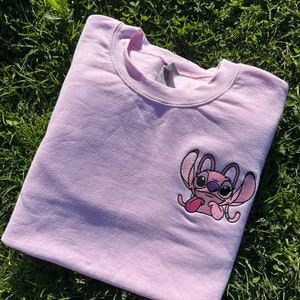 Puede incluir: Una sudadera rosa con una imagen bordada de Stitch de Lilo & Stitch. Stitch es rosa con ojos azules, saca la lengua y tiene los brazos levantados.