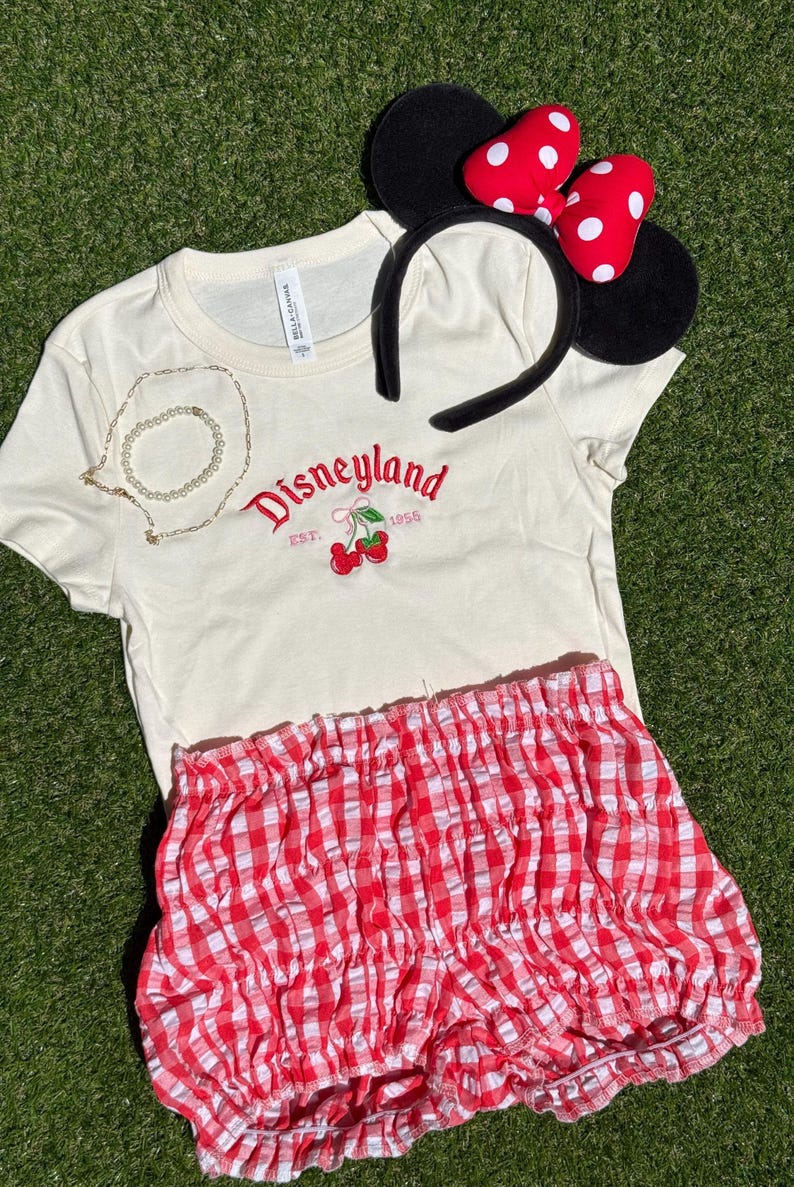Puede incluir: Una camiseta color crema con "Disneyland" en letras rojas y un gr&aacute;fico de cerezas, combinada con pantalones cortos de cuadros rojos y blancos y una diadema de orejas de Minnie Mouse con un lazo rojo y lunares blancos. Tambi&eacute;n se incluye un collar dorado.