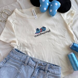 May include: An off-white t-shirt with an embroidered cartoon character and the word "overstimulated". Light blue denim shorts, a black headband with mouse ears, and a blue bow with white polka dots are also in the image.