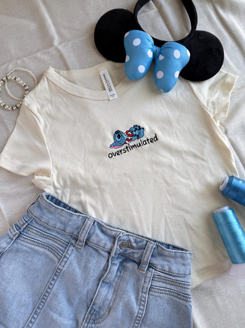 May include: An off-white t-shirt with an embroidered cartoon character and the word "overstimulated". Light blue denim shorts, a black headband with mouse ears, and a blue bow with white polka dots are also in the image.