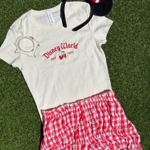 K&ouml;nnte beinhalten: Cremefarbenes T-Shirt mit "Disney World" Logo und Kirschen, kombiniert mit rot-wei&szlig; karierten Shorts. Inklusive Minnie Mouse Ohren-Haarreif mit schwarzen Ohren und roter Schleife mit wei&szlig;en Punkten. Eine goldene Halskette ist ebenfalls enthalten.