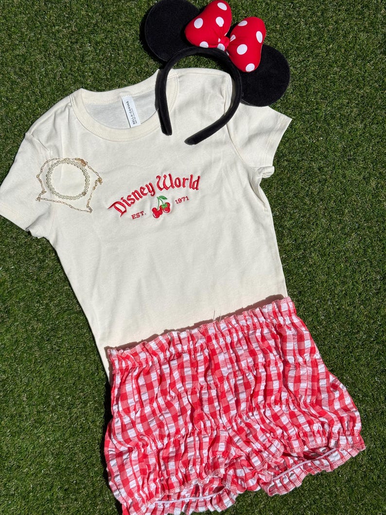 K&ouml;nnte beinhalten: Cremefarbenes T-Shirt mit "Disney World" Logo und Kirschen, kombiniert mit rot-wei&szlig; karierten Shorts. Inklusive Minnie Mouse Ohren-Haarreif mit schwarzen Ohren und roter Schleife mit wei&szlig;en Punkten. Eine goldene Halskette ist ebenfalls enthalten.