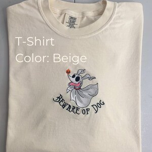 Puede incluir: Camiseta beige con una imagen bordada de Zero, el perro fantasma de Pesadilla antes de Navidad, sosteniendo una calabaza. El texto "Beware of Dog" est&aacute; bordado debajo de la imagen.