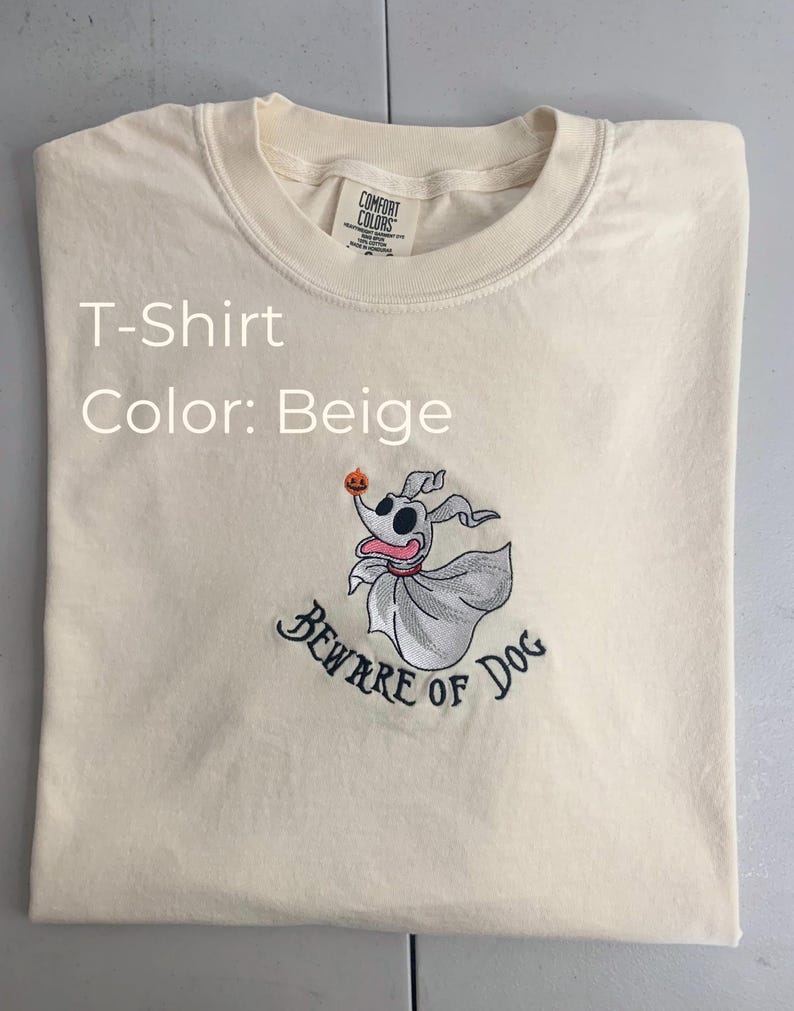 Puede incluir: Camiseta beige con una imagen bordada de Zero, el perro fantasma de Pesadilla antes de Navidad, sosteniendo una calabaza. El texto "Beware of Dog" est&aacute; bordado debajo de la imagen.