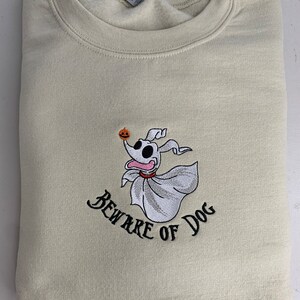 Puede incluir: Una sudadera beige con una imagen bordada de un perro fantasma sosteniendo una calabaza. El texto "Beware of Dog" est&aacute; bordado debajo de la imagen.
