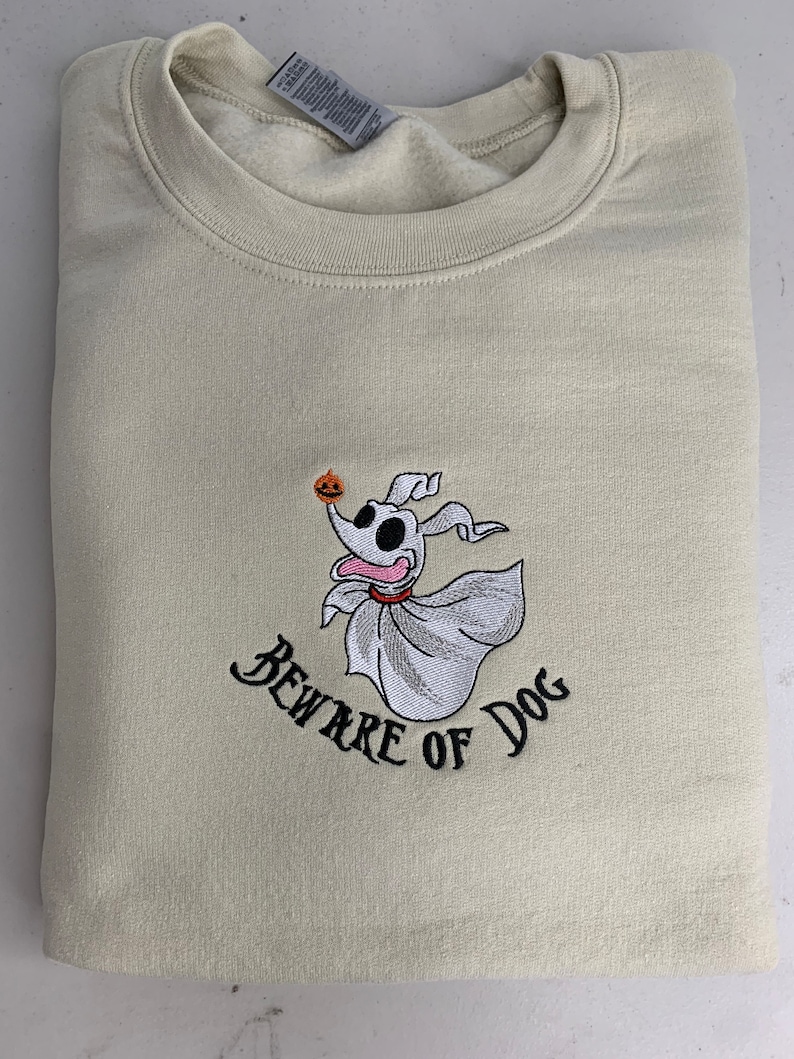 Puede incluir: Una sudadera beige con una imagen bordada de un perro fantasma sosteniendo una calabaza. El texto "Beware of Dog" est&aacute; bordado debajo de la imagen.