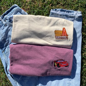May include: Two t-shirts with embroidered designs. The top shirt is beige with a "Cozy Cone Motel" sign. The bottom shirt is maroon with a red Lightning McQueen car.