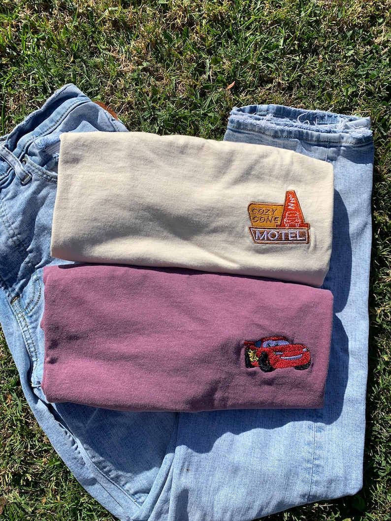 May include: Two t-shirts with embroidered designs. The top shirt is beige with a "Cozy Cone Motel" sign. The bottom shirt is maroon with a red Lightning McQueen car.