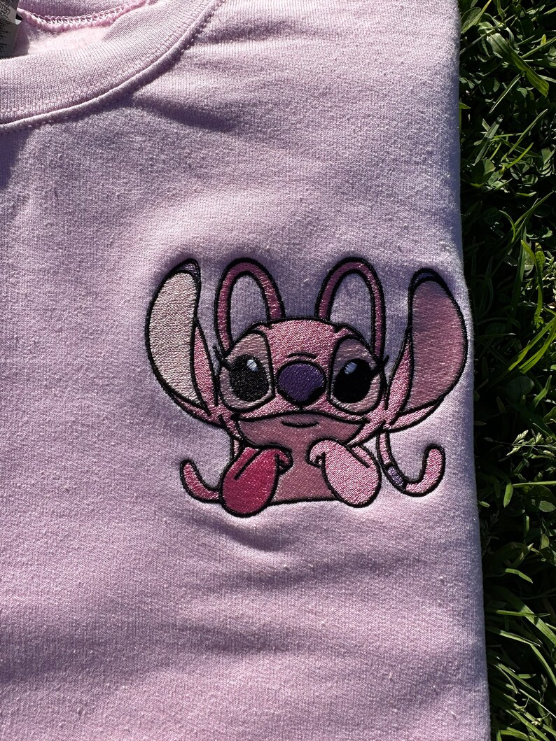 Puede incluir: Camiseta rosa con una imagen bordada de Stitch, un personaje alien&iacute;gena rosa de la pel&iacute;cula de Disney Lilo & Stitch. Stitch se muestra con la lengua fuera y los brazos cruzados.