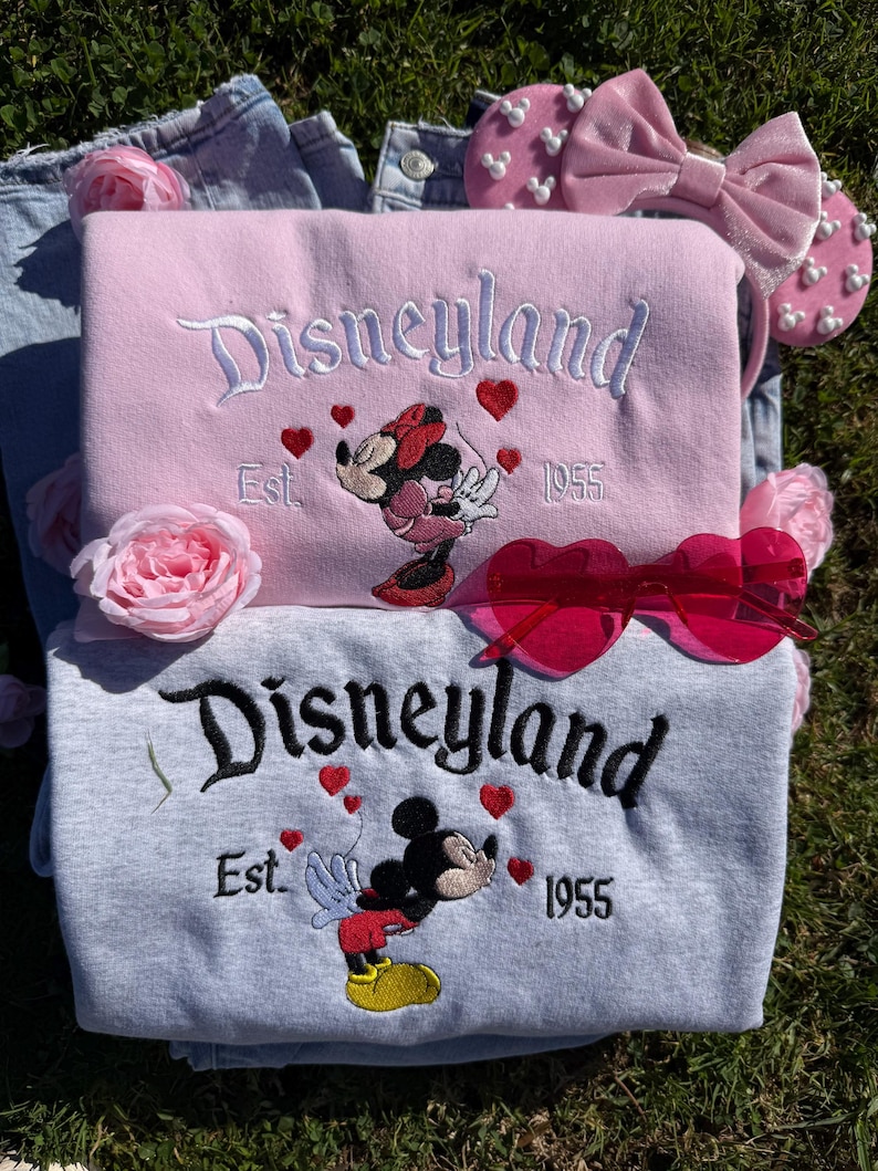 May include: Two sweatshirts, one pink and one gray, embroidered with "Disneyland Est. 1955" and a cartoon character. Pink heart accents and a Minnie Mouse design adorn the pink sweatshirt, while the gray one features Mickey Mouse. Pink heart-shaped sunglasses and a pink bow headband are also present.