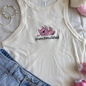 May include: Cream-colored tank top with a pink cartoon character embroidered on the front, along with the word "overstimulated" in black. Light blue denim shorts and a pearl bracelet are also in the image.