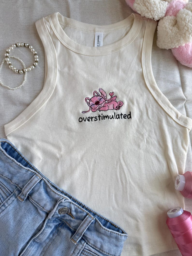 May include: Cream-colored tank top with a pink cartoon character embroidered on the front, along with the word "overstimulated" in black. Light blue denim shorts and a pearl bracelet are also in the image.