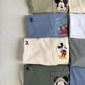 Puede incluir: Cuatro camisetas plegadas con bolsillos en el pecho con dise&ntilde;os bordados de Mickey Mouse. La camiseta 1 es verde oliva con una cara de Mickey. La 2 es azul polvoriento con Mickey aprendiz de brujo. La 3 es crema con Mickey de pie. La 4 es verde oscuro con Mickey safari.