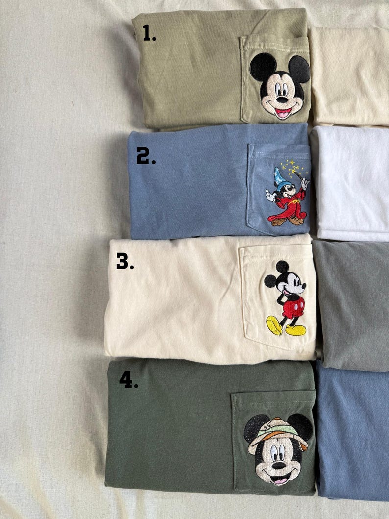 Puede incluir: Cuatro camisetas plegadas con bolsillos en el pecho con dise&ntilde;os bordados de Mickey Mouse. La camiseta 1 es verde oliva con una cara de Mickey. La 2 es azul polvoriento con Mickey aprendiz de brujo. La 3 es crema con Mickey de pie. La 4 es verde oscuro con Mickey safari.