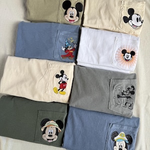 Puede incluir: Una colecci&oacute;n de ocho camisetas dobladas en varios colores apagados, cada una con un parche bordado &uacute;nico de Mickey Mouse en el bolsillo del pecho. Los dise&ntilde;os incluyen a Mickey con diferentes atuendos, como mago, marinero y explorador.