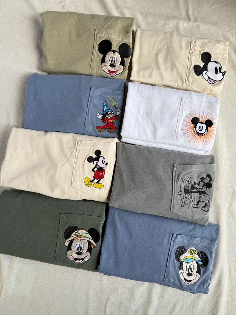 Puede incluir: Una colecci&oacute;n de ocho camisetas dobladas en varios colores apagados, cada una con un parche bordado &uacute;nico de Mickey Mouse en el bolsillo del pecho. Los dise&ntilde;os incluyen a Mickey con diferentes atuendos, como mago, marinero y explorador.