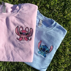 Puede incluir: Dos camisetas con personajes de Stitch bordados. La camiseta de la izquierda es rosa con un personaje de Stitch rosa. La camiseta de la derecha es azul claro con un personaje de Stitch azul.