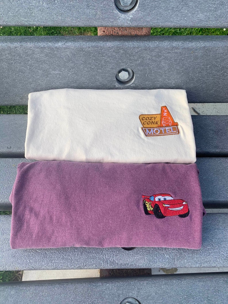 May include: Two t-shirts, one white and one maroon, with embroidered designs. The white t-shirt has a motel sign with the text "Cozy Cone Motel". The maroon t-shirt has a red cartoon race car.