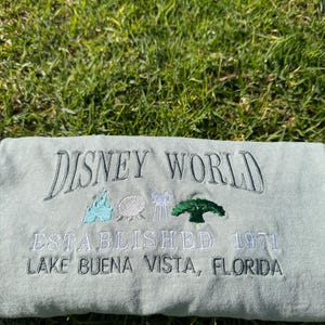 Puede incluir: Una camiseta verde claro con el texto "DISNEY WORLD ESTABLISHED 1971 LAKE BUENA VISTA, FLORIDA" bordado en negro. La camiseta presenta un peque&ntilde;o dise&ntilde;o bordado del castillo de Disney World, una nave espacial, un paseo en taza de t&eacute; y un &aacute;rbol.