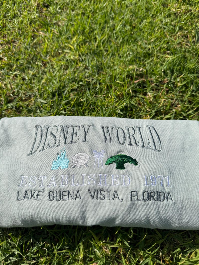 Puede incluir: Una camiseta verde claro con el texto "DISNEY WORLD ESTABLISHED 1971 LAKE BUENA VISTA, FLORIDA" bordado en negro. La camiseta presenta un peque&ntilde;o dise&ntilde;o bordado del castillo de Disney World, una nave espacial, un paseo en taza de t&eacute; y un &aacute;rbol.