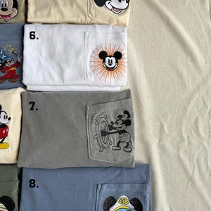 Puede incluir: Una colecci&oacute;n de cuatro camisetas plegadas con bolsillos en el pecho, numeradas del 5 al 8. La camiseta 5 es crema con una cabeza de Mickey Mouse. La 6 es blanca con un dise&ntilde;o de Mickey y rayos de sol. La 7 es gris con Mickey al tim&oacute;n. La 8 es azul con Mickey capit&aacute;n.
