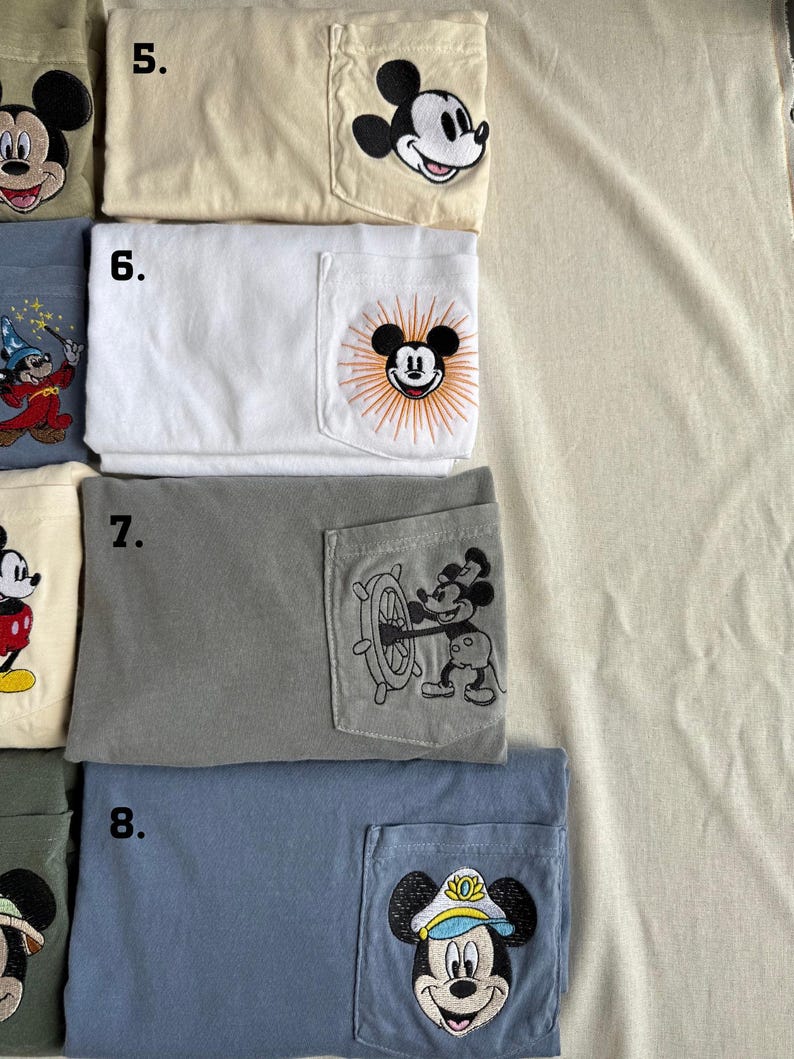 Puede incluir: Una colecci&oacute;n de cuatro camisetas plegadas con bolsillos en el pecho, numeradas del 5 al 8. La camiseta 5 es crema con una cabeza de Mickey Mouse. La 6 es blanca con un dise&ntilde;o de Mickey y rayos de sol. La 7 es gris con Mickey al tim&oacute;n. La 8 es azul con Mickey capit&aacute;n.