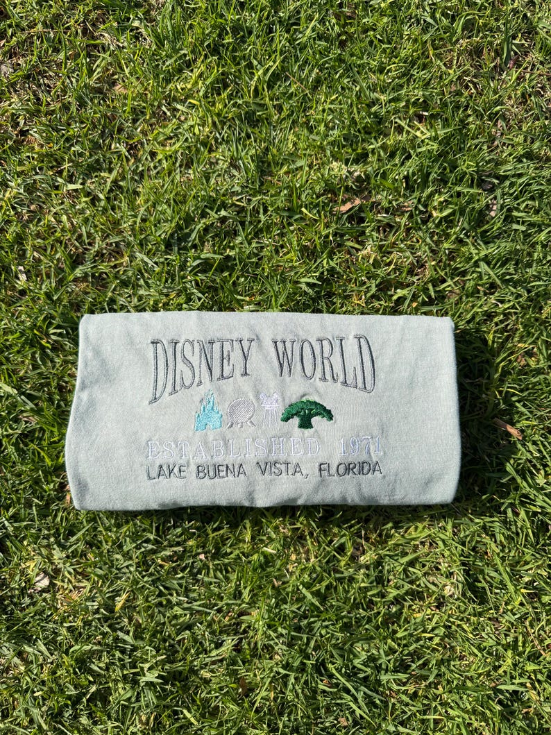 Puede incluir: Una camiseta azul claro con el texto "Disney World Established 1971 Lake Buena Vista, Florida" bordado en negro. La camiseta tambi&eacute;n presenta im&aacute;genes bordadas del castillo de Disney World, Spaceship Earth y el &Aacute;rbol de la Vida.