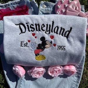 May include: A gray sweatshirt with the word "Disneyland" embroidered in black script, along with "Est. 1955" and a Mickey Mouse design. The sweatshirt is laid on top of a pair of light blue jeans, with pink flowers and a pink bow.