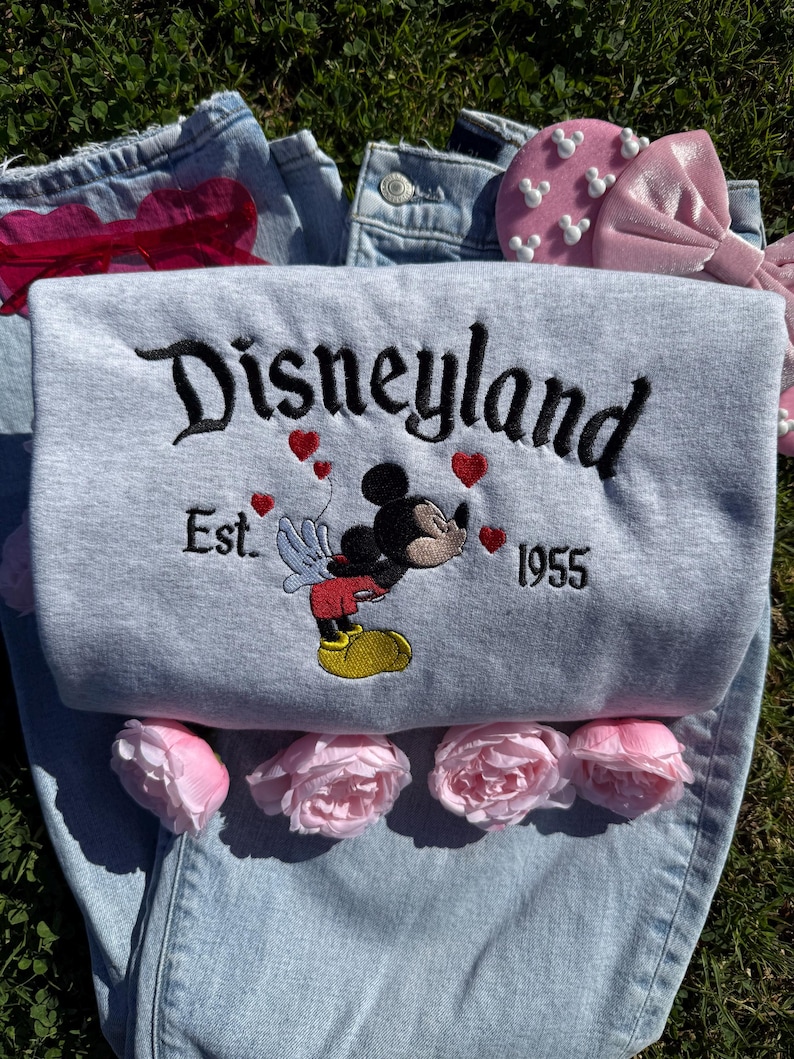 May include: A gray sweatshirt with the word "Disneyland" embroidered in black script, along with "Est. 1955" and a Mickey Mouse design. The sweatshirt is laid on top of a pair of light blue jeans, with pink flowers and a pink bow.