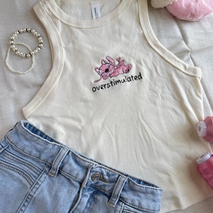 May include: Cream-colored tank top with a pink cartoon character and the word "overstimulated" embroidered in black. Light blue denim shorts are visible below the top. A pearl bracelet and pink slippers are also in the image.