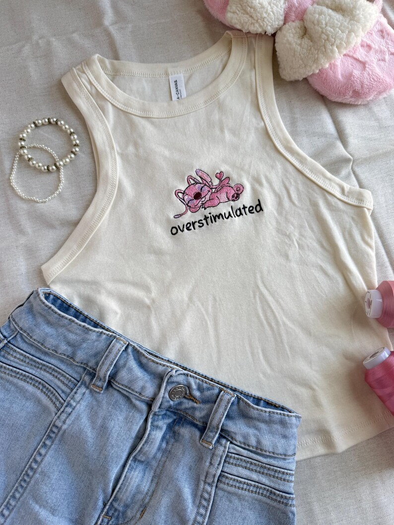 May include: Cream-colored tank top with a pink cartoon character and the word "overstimulated" embroidered in black. Light blue denim shorts are visible below the top. A pearl bracelet and pink slippers are also in the image.