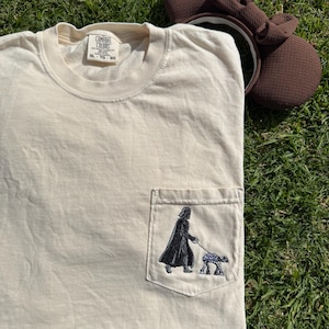 May include: A beige t-shirt with a pocket featuring an embroidered image of Darth Vader walking a small droid. The t-shirt has a tag that reads "Comfort Colors" and "XL - TG - EG".