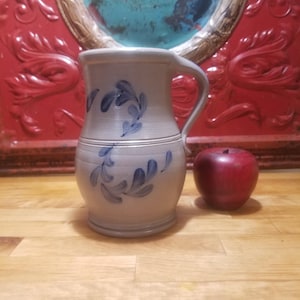 May include: A stoneware pitcher with a handle, decorated with blue floral patterns. The pitcher is light gray with a dark blue design. A red apple sits next to the pitcher. The pitcher is a decorative item.